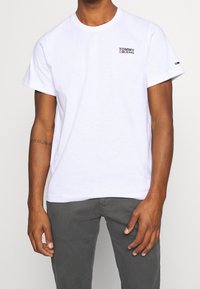 Vit bomullst-shirt med en liten svart och röd "TOMMY JEANS"-logotyp på vänster bröst och korta ärmar. Texturerat tyg med rund hals.