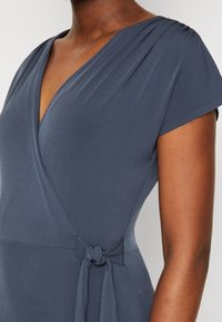 Nahaufnahme einer dunkelhäutigen Frau, die ein graues Wickelkleid mit kurzen Ärmeln trägt, das an der Taille mit einer seitlichen Schleife gebunden ist.