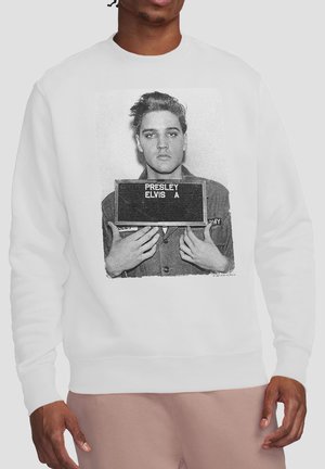 Man wearing white sweatshirt with black and white print of Elvis Presley holding a sign with his name in a military uniform.