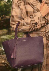 Sac fourre-tout en cuir violet avec une finition texturée, doté de deux poignées et d'un gland décoratif. Logo argenté à l'avant.