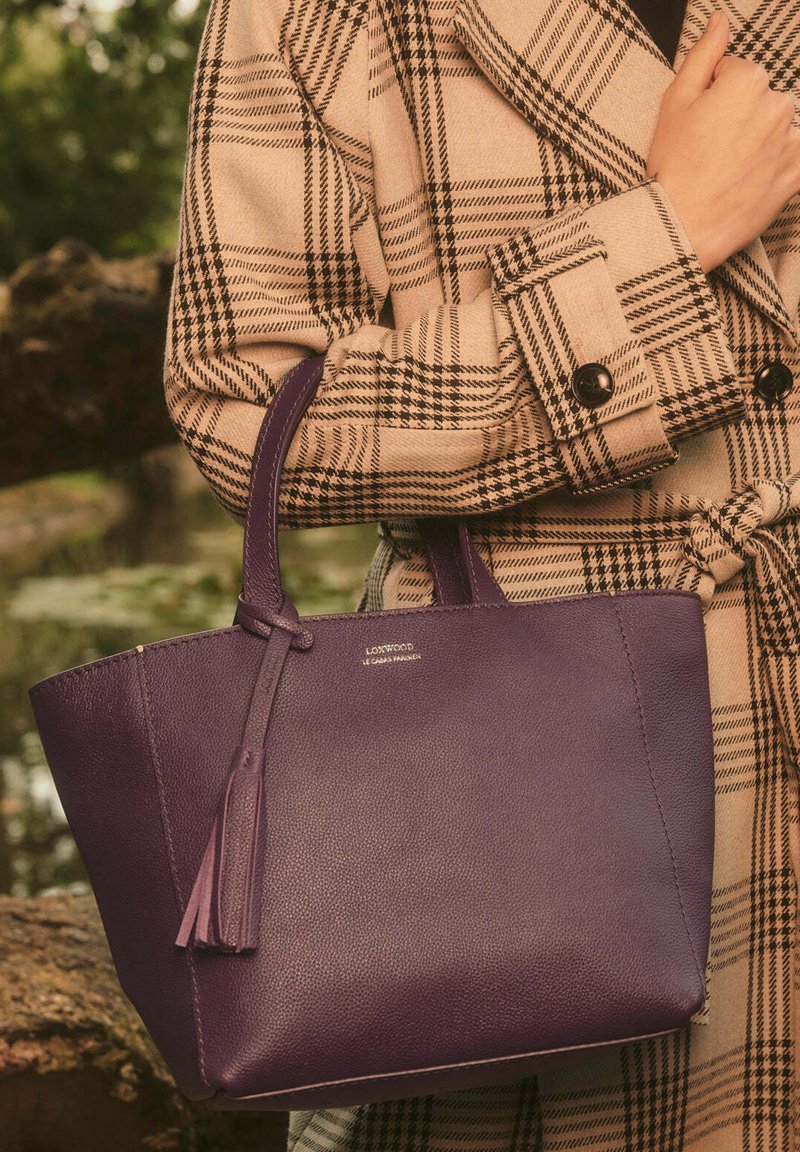 Sac fourre-tout en cuir violet avec une finition texturée, doté de deux poignées et d'un gland décoratif. Logo argenté à l'avant.