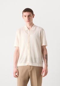 Chemise en tricot crème à manches courtes avec des rayures verticales, fermeture à boutons et une texture subtile ; associée à un pantalon beige.