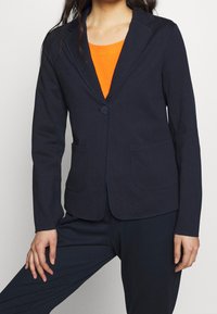 Donna che indossa un blazer blu navy allacciato una volta sopra una maglia arancione, abbinato a pantaloni blu navy, in piedi di fronte a uno sfondo chiaro uniforme.