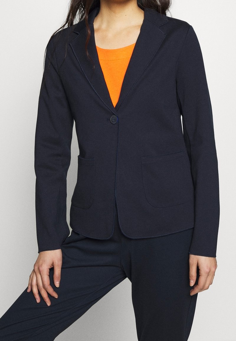 Donna che indossa un blazer blu navy allacciato una volta sopra una maglia arancione, abbinato a pantaloni blu navy, in piedi di fronte a uno sfondo chiaro uniforme.