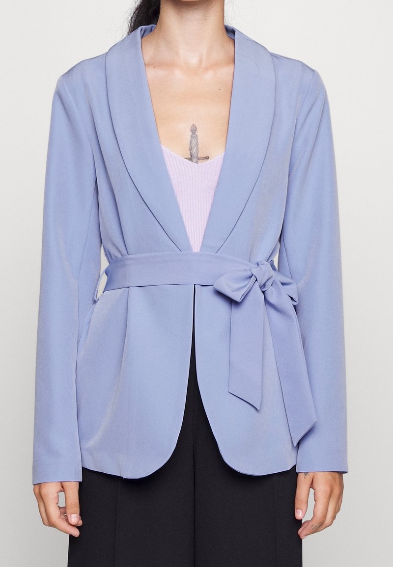 Personne portant un blazer violet clair avec une ceinture nouée sur un haut lavande, révélant un tatouage de dague sur le haut de la poitrine.