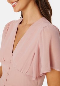 Blouse rose clair avec des manches bouffantes, un décolleté en V profond et des boutons décoratifs sur le devant, réalisée dans un tissu doux et aéré.
