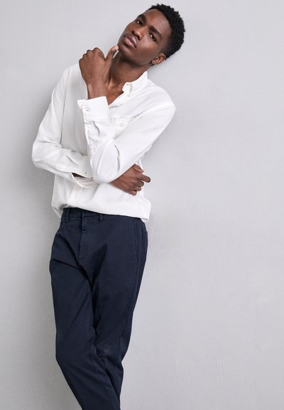 Camisa blanca de manga larga con botones, combinada con pantalones azul marino. La tela parece suave; la camisa tiene un ajuste ligeramente holgado.