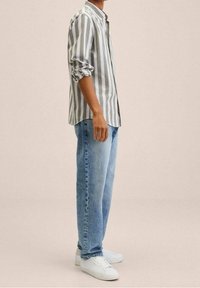 Camicia a righe grigia chiaro e bianca, abbinata a jeans blu chiaro e sneaker bianche, esposta in una posa in piedi.