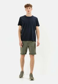 Navy cotton t-shirt paired with rolled olive green shorts. Model wears gray sneakers with orange accents, standing against a white background.