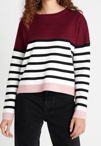 Sweter w paski w kolorach bordowym, czarnym i białym z różowym obszyciem, wykonany z miękkiego dzianinowego materiału; posiada okrągły dekolt i długie rękawy.