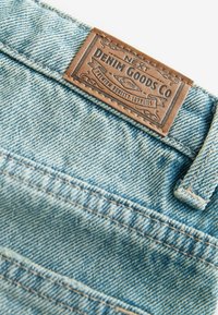 Hellblaues Denim-Gewebe mit einem braunen Lederschild, auf dem "NEXT DENIM GOODS CO." steht. Das Schild hat dekorative Nähte und eine strukturierte Oberfläche.