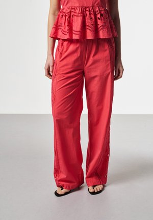 Pantalon rouge à jambes larges avec motifs en dentelle découpés sur les côtés, associé à un haut péplum assorti et des sandales noires à bouts ouverts sur un sol clair.