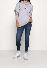 Felpa grigia con cappuccio e lacci, con logo "TOMMY JEANS". Abbinata a jeans skinny blu scuro con risvolto e sneakers bianche.
