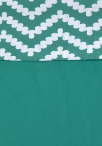 Tessuto lavorato a maglia color teal con una sezione piegata che mostra motivi a zigzag e croce bianchi su sfondo teal.