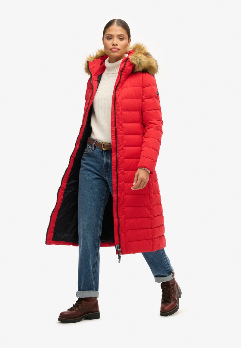 Rood gewatteerd lang jasje met een capuchon met bontafwerking, ritssluiting en contrasterende zwarte voering, gecombineerd met lichtblauwe jeans en bruin enkellaarsjes.