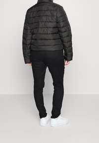 Přefukovací bunda v tmavě černé barvě s prošívaným designem, spárovaná s černými slim fit džínami a bílými teniskami. Zadní pohled, který zdůrazňuje texturu a tvar.