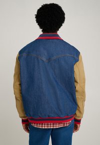 Denim and beige jacket with red ribbed collar and cuffs, featuring contrasting sleeves and a curved seam design. Worn over a plaid shirt.