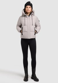 Lys grå pufferjakke med hætte, med sorte accenter, to frontlommer og justerbare snore. Kombineret med sorte leggings og støvler.
