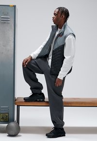 Gray quilted vest over a white long-sleeve shirt, paired with gray sweatpants and black athletic shoes. Setting includes a bench and locker.