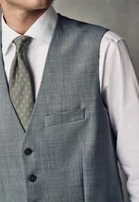 Chaleco de tela gris con una textura suave, que incluye un bolsillo en el pecho, dos botones y una corbata de patrón verde oliva sobre una camisa blanca.