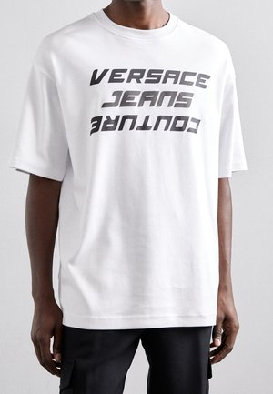 Person trägt ein lockeres weißes T-Shirt mit schwarzem fettem Text "VERSACE JEANS COUTURE" auf der Vorderseite und schwarze Hose.