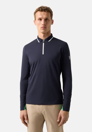 Mand iført marineblå poloshirt med langærmer og hvid lynlås, beige bukser, stående foran en ensfarvet lys grå baggrund.