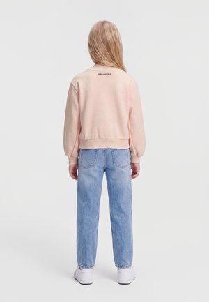 Barn med langt blond hår iført lyserød sweatshirt, blå jeans og hvide sneakers, der står med ryggen til på en ensfarvet baggrund.