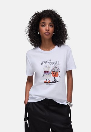 Donna con capelli ricci che indossa una maglietta bianca con una vignetta di un cartone di latte e una cupcake che si tengono per mano, con la scritta "Coppia Perfetta".