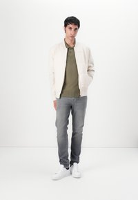 Hellbeige Bomberjacke mit glatter Textur, olivgrünes Poloshirt, graue Jeans und weiße Sneaker. Einfaches, lässiges Outfit.