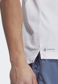 Camisa atlética blanca con un panel lateral texturizado, que presenta la marca "AEROREADY" y un corte elegante. Combinada con pantalones cortos gris claro.