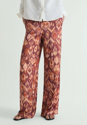 Persona que lleva pantalones anchos y sueltos con un patrón geométrico en tonos óxido y beige, combinados con una camisa blanca abotonada y sandalias negras.