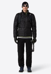 Homme portant une doudoune noire, des gants noirs, un pantalon noir, un bonnet, des lunettes de soleil, des bottes de randonnée bleues et portant un petit sac à dos noir avec des sangles de poitrine.