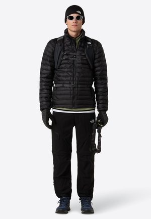 Uomo che indossa una giacca a vento nera, guanti neri, pantaloni neri, berretto, occhiali da sole, scarponi da trekking blu e porta uno zaino nero di piccole dimensioni con cinghie sul petto.