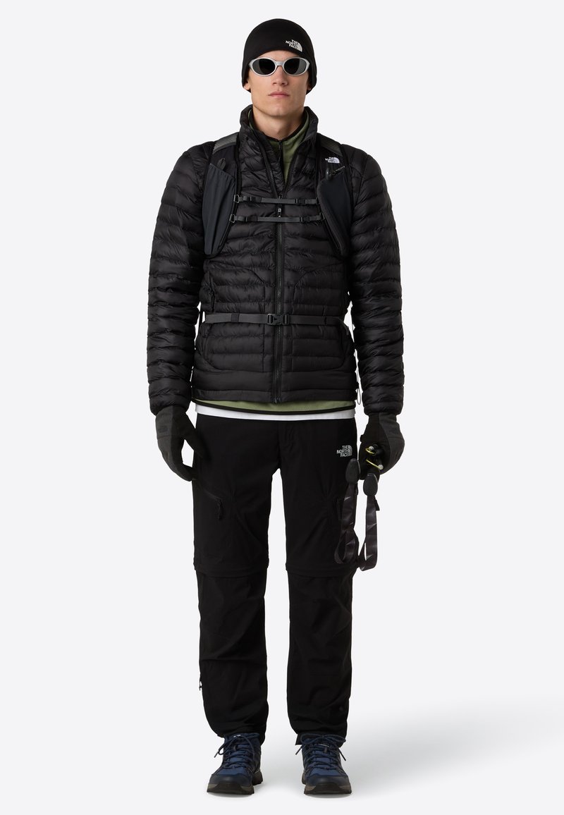 Uomo che indossa una giacca a vento nera, guanti neri, pantaloni neri, berretto, occhiali da sole, scarponi da trekking blu e porta uno zaino nero di piccole dimensioni con cinghie sul petto.