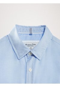  Lichtblauwe geklede shirt met twee witte knopen vastgemaakt, met een kleine stoflus en merklabel zichtbaar binnen de kraag.