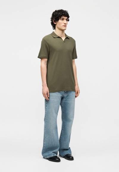 Jeune homme debout, portant un polo manches courtes vert olive, un jean large bleu clair et des chaussures noires sur un fond uni.