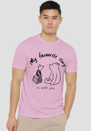 Junger Mann trägt ein rosa T-Shirt mit einer Zeichnung eines Bären und eines Ferkels, die auf Büchern sitzen, und dem Text "My favourite story is with you."