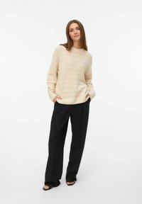 Pull en tricot beige avec un motif texturé, encolure ronde et manches longues, associé à un pantalon noir à jambes larges et des sandales.