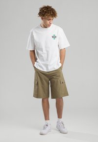 Valkoinen puuvillapaita, jossa on vihreä logo, yhdistettynä khakinvärisiin cargo-shortseihin, joissa on sivutaskut. Mallilla on valkoiset lenkkarit.