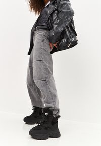 Chaqueta de cuero negra con detalles gráficos; jeans grises holgados; zapatillas negras gruesas con tela texturizada y diseño de cordones.