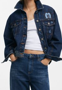 Denim takki tummansinisellä värillä, kontrastiväriset oranssit ompeleet, kaksi rintataskua ja brodeerattu Mikki Hiiri -yksityiskohta olkapäällä.
