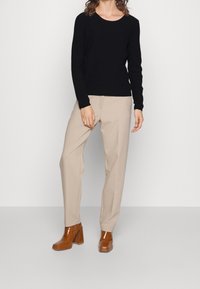 Frau trägt einen schwarzen langen Pullover, beige gerade geschnittene Hose und braune hochhackige Stiefeletten und steht vor einem neutralen Hintergrund.