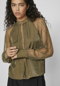 Blouse vert olive avec des manches longues en dentelle travaillée et un col montant. Dispose d'une fermeture à l'avant et d'un léger éclat dans la texture du tissu.