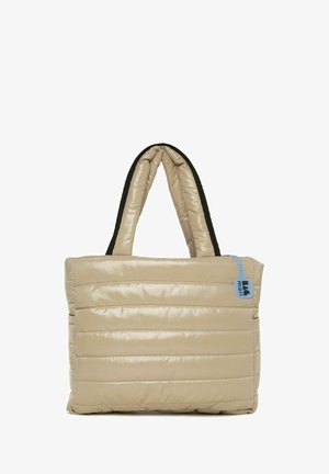 Bežs pildīts iepirkumu soma ar horizontālu quilted tekstūru, melnām rokturiem un zilu etiķeti sānos. Iespējota taisnstūra forma.