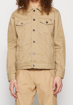 Person med krøllet hår iført en beige jakke med knapper og matchende beige bukser, stående mod en ensfarvet baggrund.