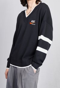Person wearing a black Nike sportswear sweater with white stripes on one sleeve and gray pleated pants, one hand in pocket.