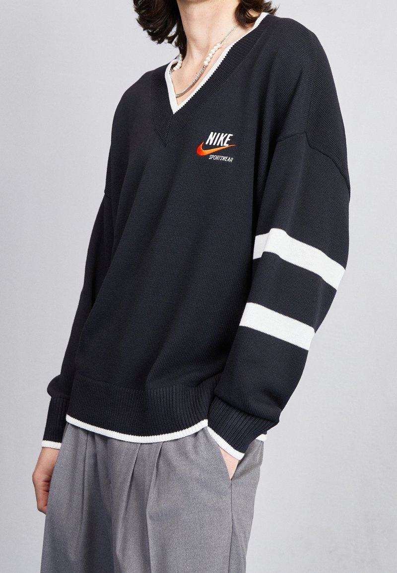 Person wearing a black Nike sportswear sweater with white stripes on one sleeve and gray pleated pants, one hand in pocket.