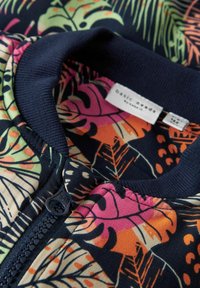 Veste zippée avec un col côtelé bleu marine ; présente un imprimé tropical coloré de feuilles en rose, vert et orange sur un fond sombre.