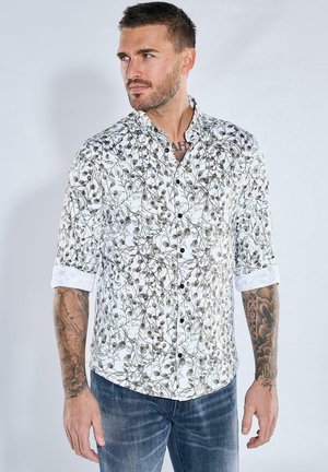 Weißes Button-Down-Hemd mit einem blumigen Muster in Grün und Braun, mit hochgerollten Ärmeln und einem Kragen. Modell trägt Jeans.