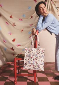 Moteris šviesiai mėlynu aprangos komplektu remiasi į raudoną medinį kėdę, laikydama smėlio spalvos gėlių rašto nešiojamą krepšį priešais žvaigždėmis margintą smėlio spalvos foną.